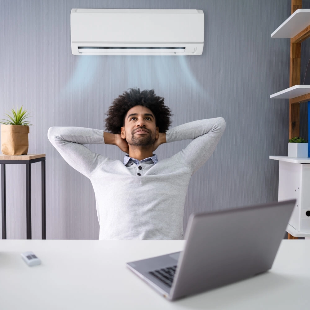 Homem com uma expressão feliz sentado de frente para um notebook e atrás dele um ar condicionado